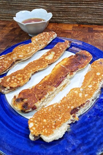 Four long pancake-battered bacon strips on a blue plate with a small bowl of syrup.