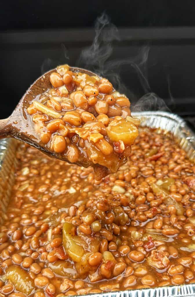 Steaming wooden spoon lifting apple pie baked beans from a foil tray.