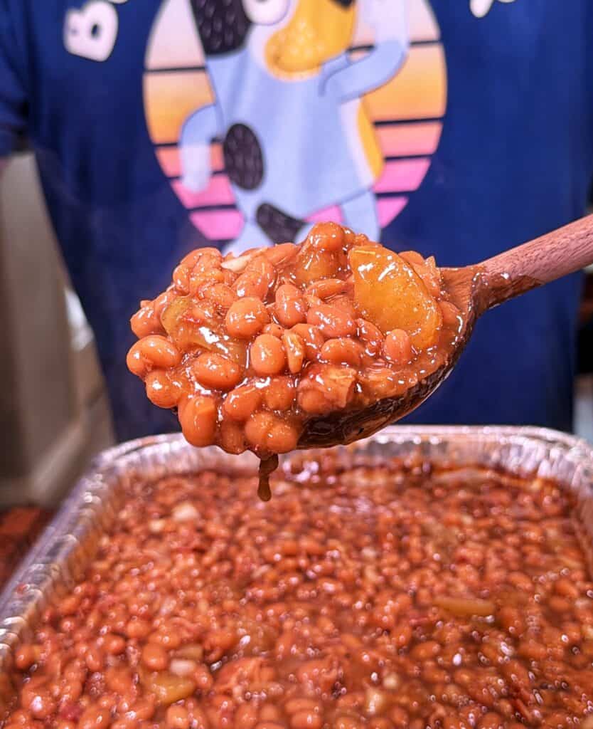 Wooden spoon scooping apple pie baked beans from a foil tray.