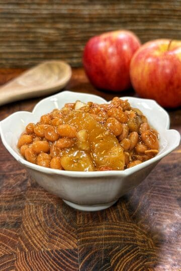 White bowl of apple pie baked beans on a wooden surface with red apples and a wooden spoon in the background.
