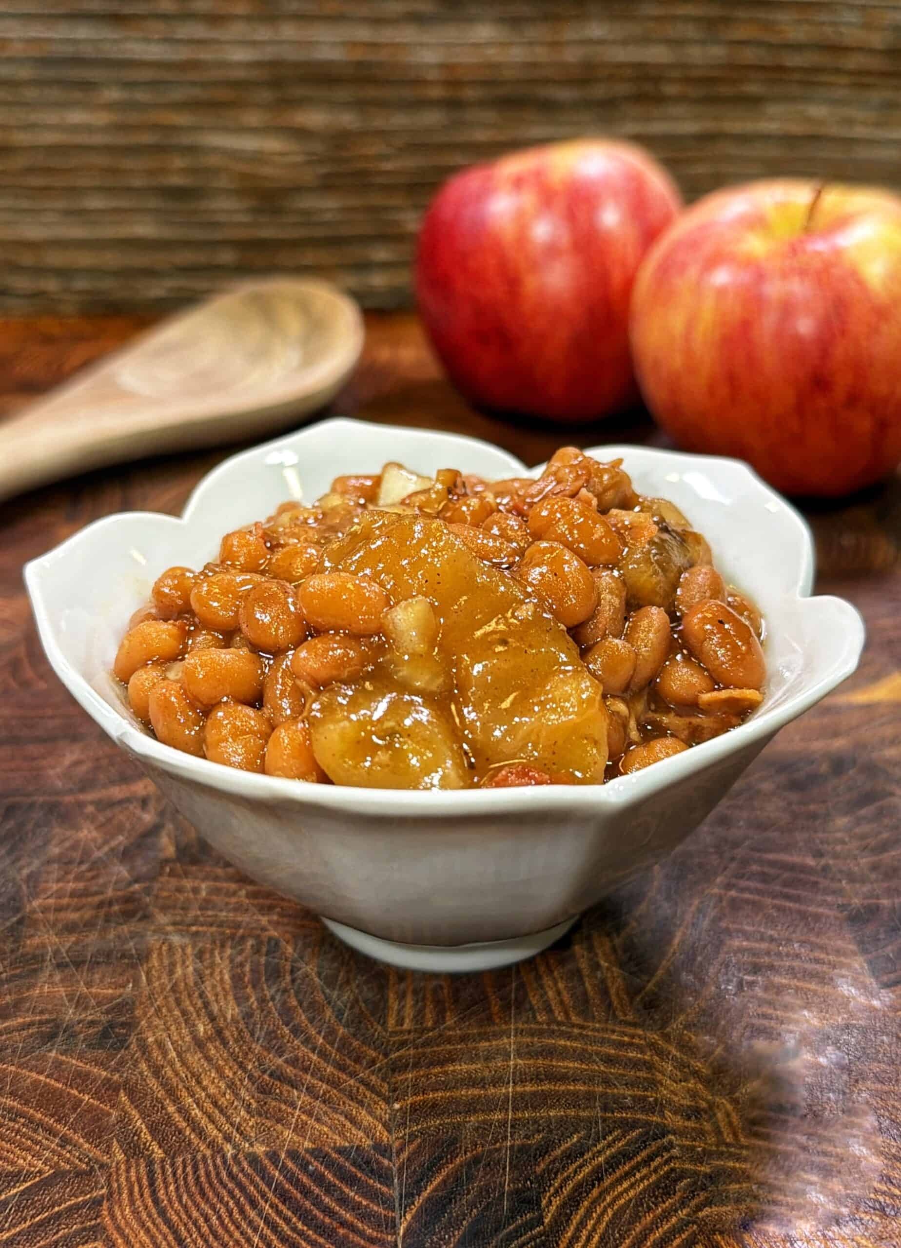 White bowl of apple pie baked beans on a wooden surface with red apples and a wooden spoon in the background.