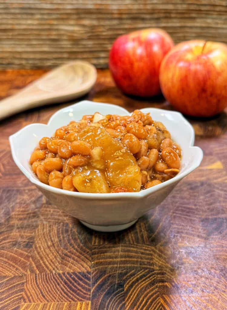 White bowl of apple pie baked beans with apple chunks on a wooden surface with red apples and a wooden spoon in the background.