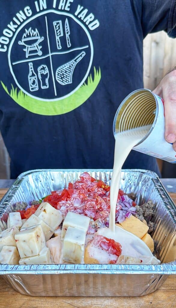 A person pours liquid from a can into a foil pan filled with cubed cheese, diced tomatoes, and onions for smoked cheeseburger queso.