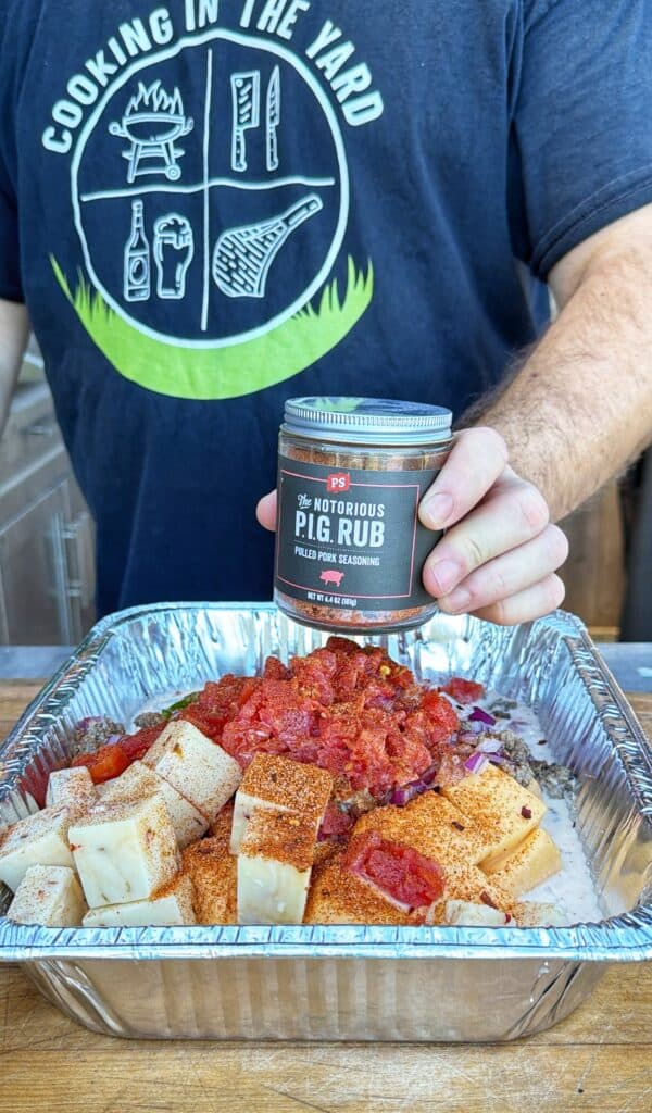 A jar of seasoning is held over a foil pan filled with cheese, diced tomatoes, and ground beef for smoked cheeseburger queso.