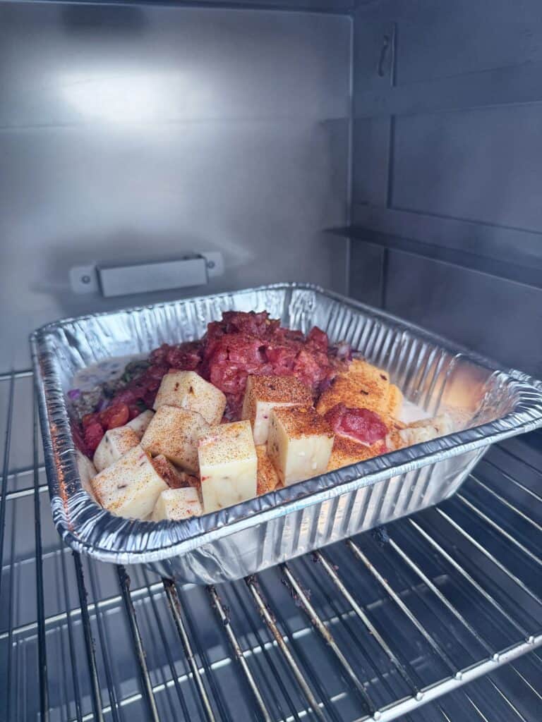 A pan of cubed cheese, ground beef, and diced vegetables for smoked cheeseburger queso sits on a smoker rack.