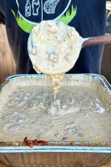 A wooden spoon lifts chunky smoked cheeseburger queso from a foil pan, with melted cheese stretching back to the pan.