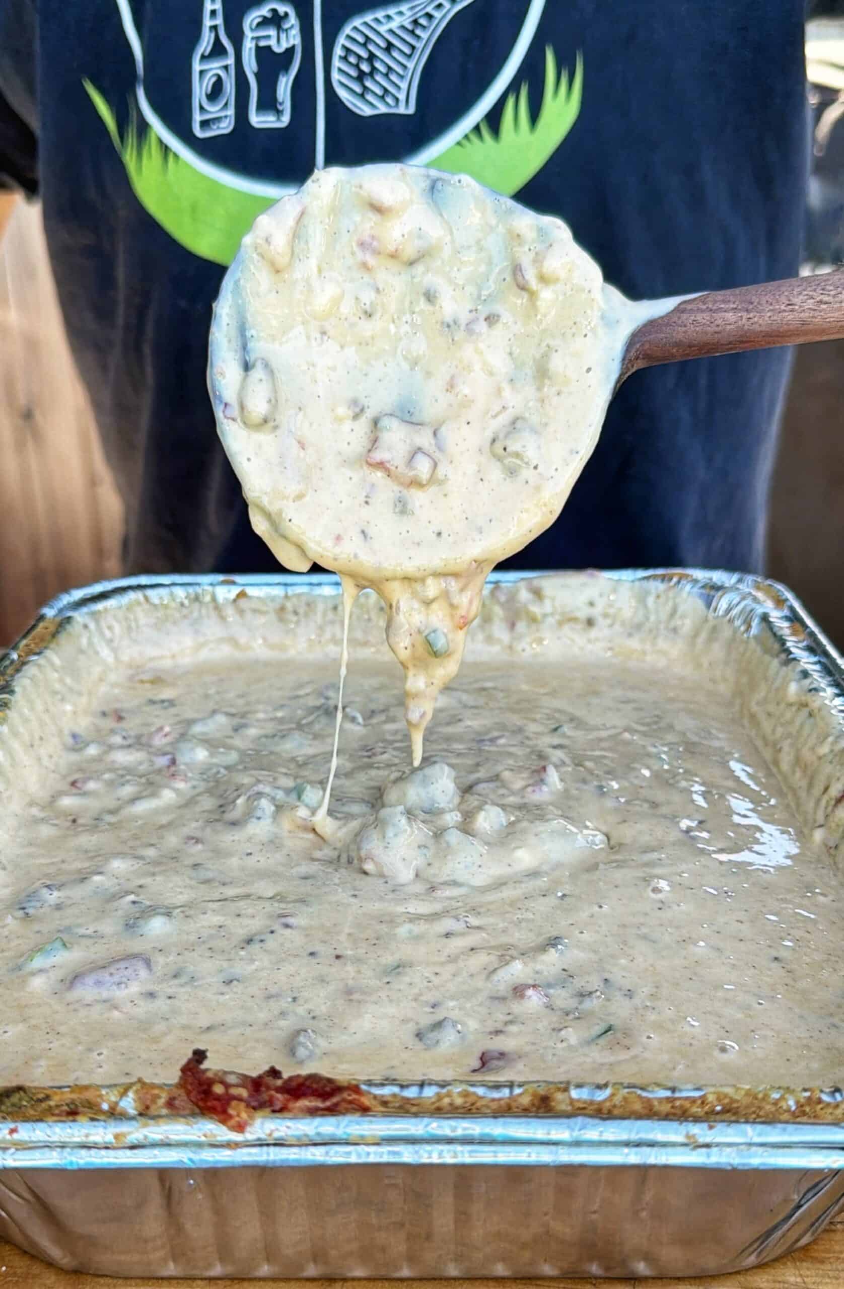 A wooden spoon lifts chunky smoked cheeseburger queso from a foil pan, with melted cheese stretching back to the pan.