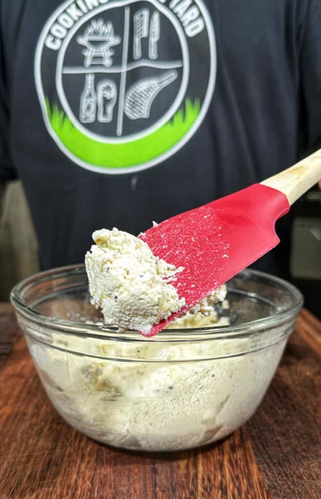 Red silicone spatula lifting seasoned cheese filling from a glass bowl.