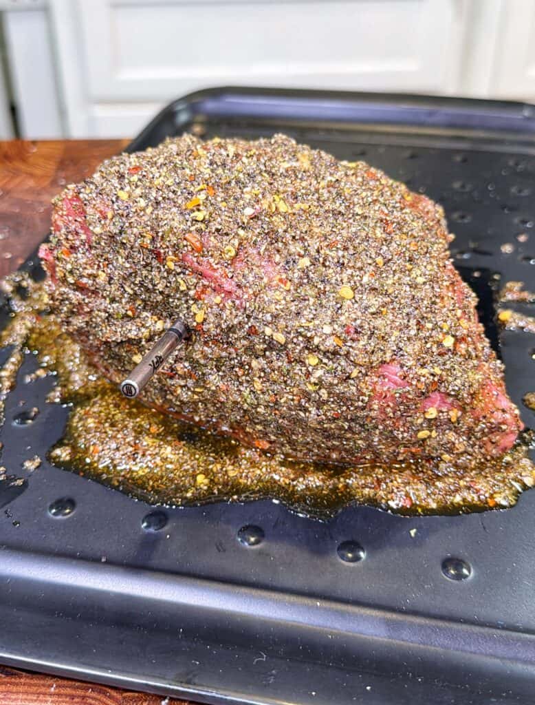 Raw beef roast covered in coarse spice rub with a meat thermometer inserted on a baking sheet.