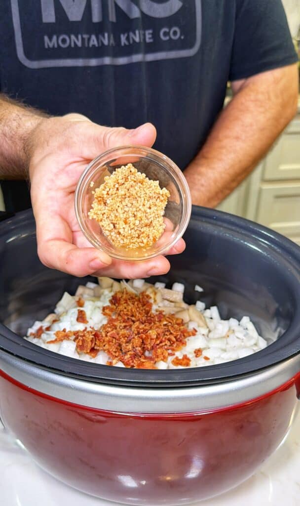 A hand holds a small glass bowl of minced garlic over a slow cooker containing chopped onions and cooked bacon.