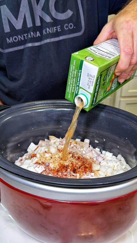 A person pours chicken broth from a carton into a slow cooker containing chopped onions and seasonings.