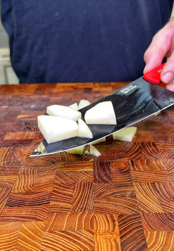 A chef's knife holds chopped potato pieces above a wooden cutting board.