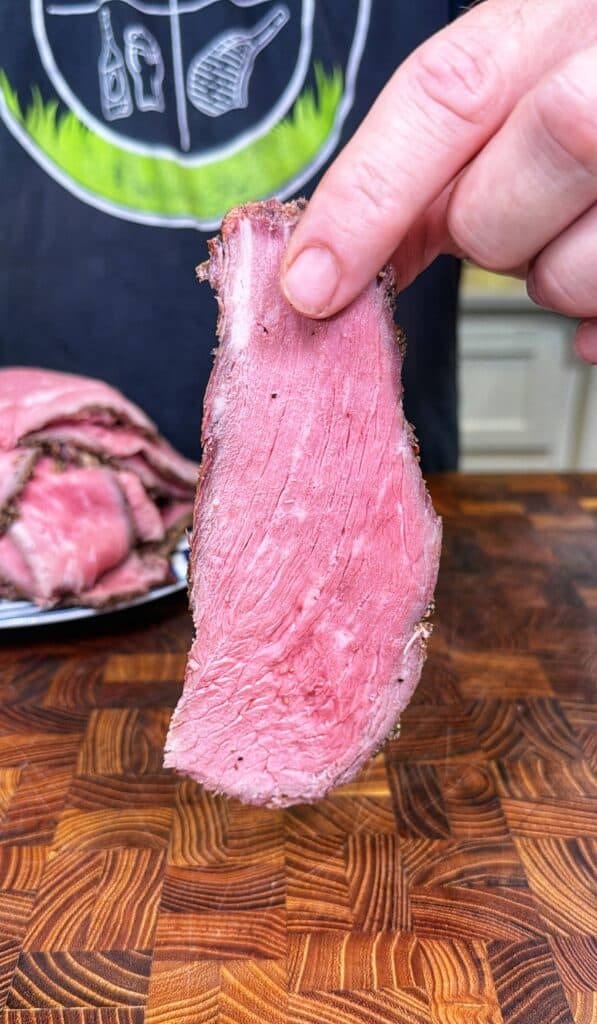 Hand holding a thin slice of deli-style roast beef above a wooden cutting board.
