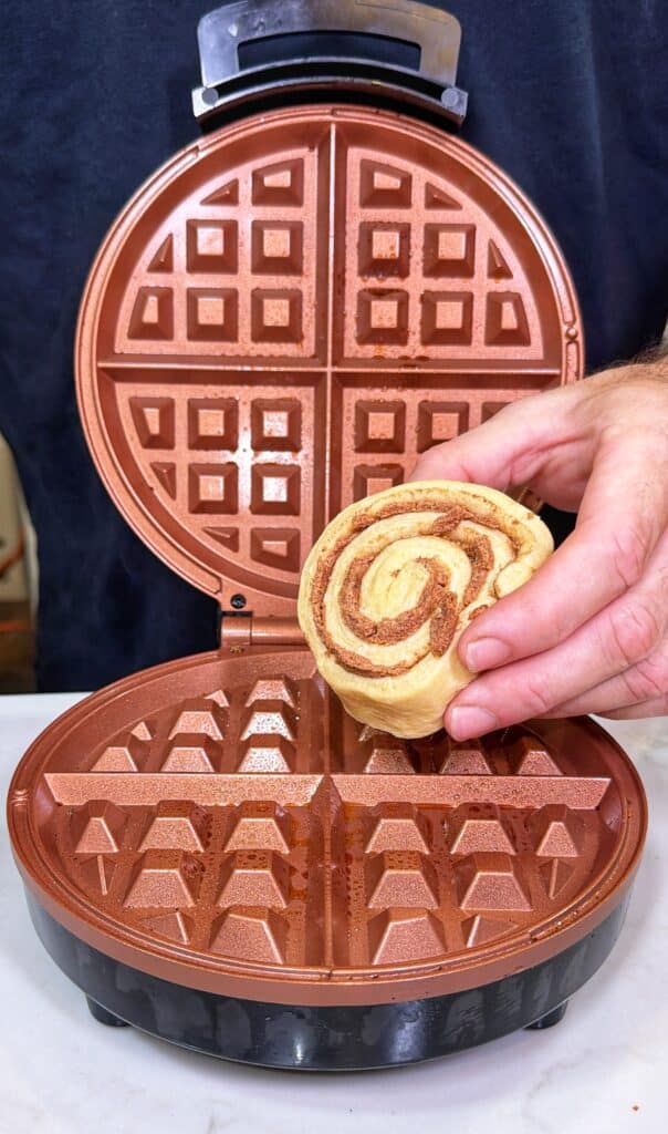 A hand places a cinnamon roll into an open waffle iron.