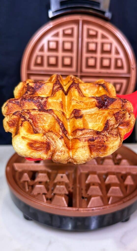 A cinnamon roll waffle being lifted from an open waffle maker with a spatula.