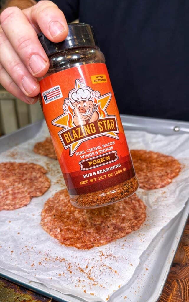 A hand sprinkles seasoning onto sausage patties on a parchment-lined baking sheet.