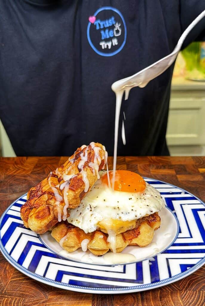A knife drizzles frosting over a cinnamon roll waffle breakfast sandwich topped with a fried egg.