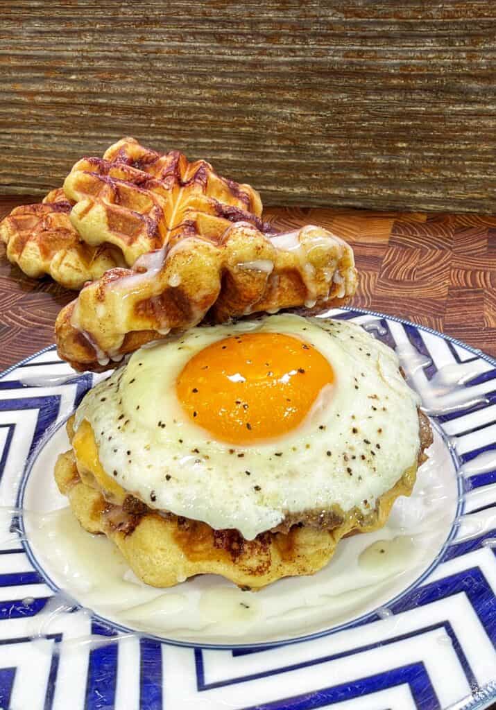 A cinnamon roll waffle breakfast sandwich with a sausage patty and sunny-side-up egg, drizzled with frosting on a patterned plate.