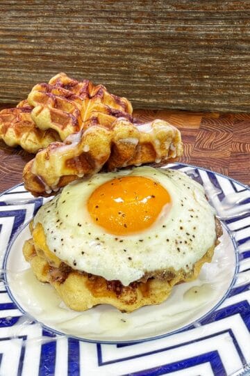 A cinnamon roll waffle breakfast sandwich topped with a sunny-side-up egg and drizzled with frosting on a patterned plate.