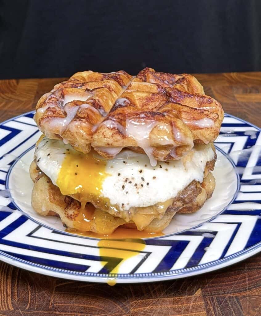 A cinnamon roll waffle breakfast sandwich with a fried egg and melted cheese, with egg yolk running onto a patterned plate.