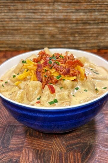 A blue bowl of loaded potato soup topped with shredded cheddar cheese, crumbled bacon, and sliced green onions.