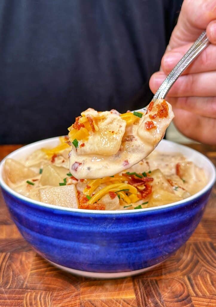 A spoon lifts loaded potato soup topped with shredded cheddar cheese, bacon, and sliced green onions from a blue bowl.
