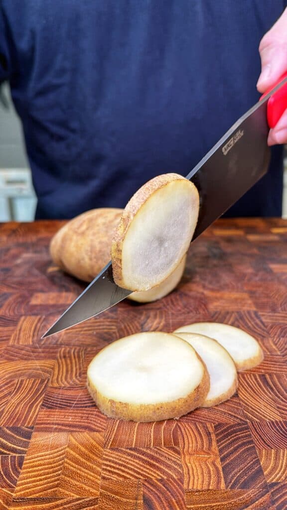 A chef's knife holds a potato slice above more potato slices on a wooden cutting board.