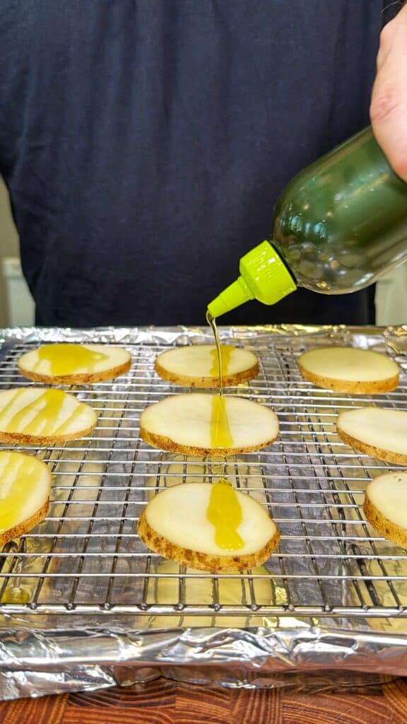 Oil being drizzled onto potato slices arranged on a wire rack.