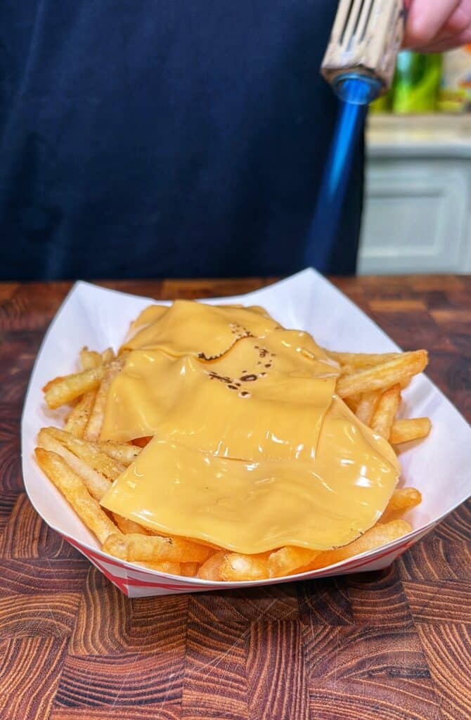 A paper tray of Cajun steak fries topped with melted cheese as a handheld torch is held over them.
