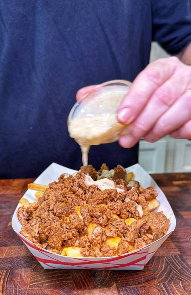 A hand pours sauce from a small container over Cajun steak fries topped with melted cheese and steak in a paper tray.