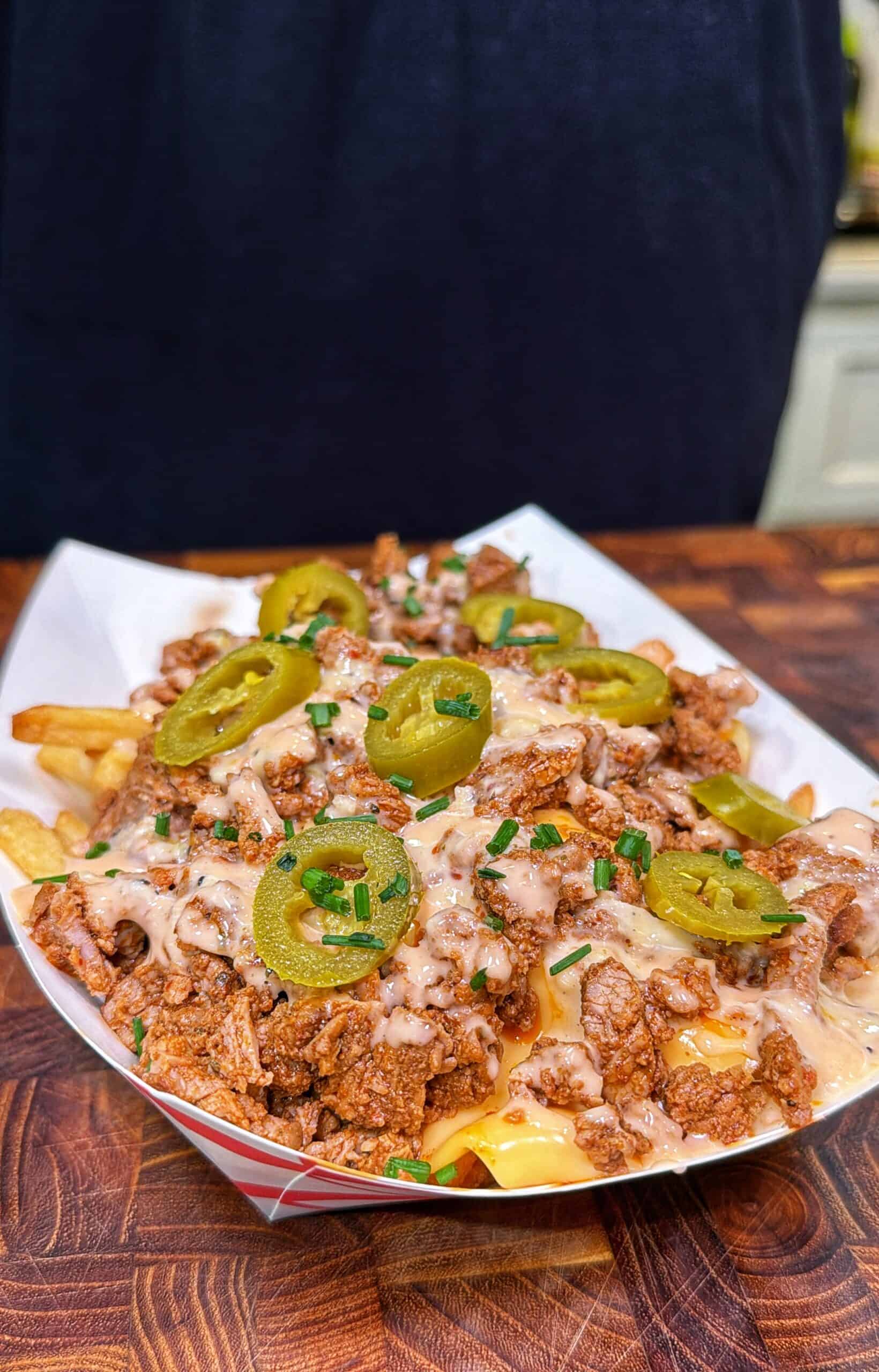 Cajun steak fries topped with steak, queso, spicy ranch, sliced jalapeños, and chives in a paper tray.