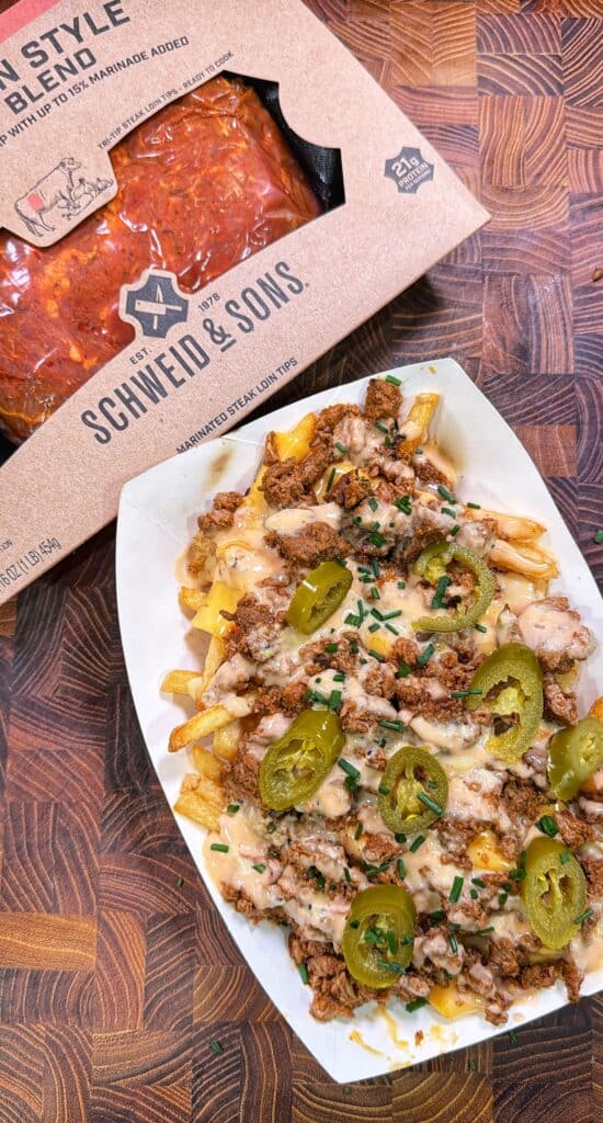 A paper tray of Cajun steak fries topped with steak, melted cheese, sliced jalapeños, and chives beside a package of Schweid & Sons marinated steak loin tips.