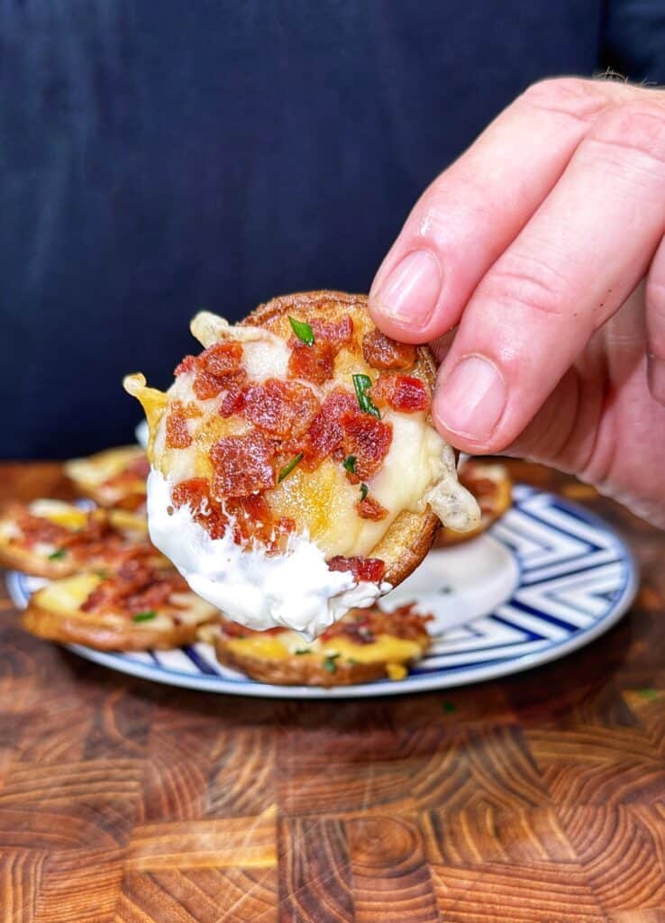 A hand holds a potato nacho topped with melted cheese, bacon, and sour cream, with more potato nachos in the background.
